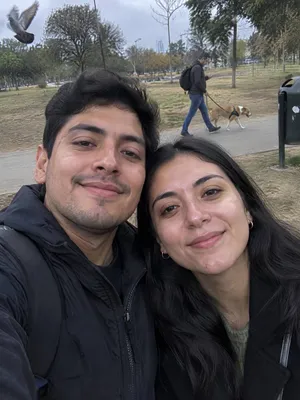 Selfie de una pareja en un parque; detrás pasan un chico con su perro y una paloma, y hay un basurero